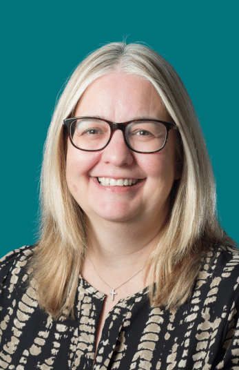 A woman with straight, shoulder-length blonde hair and glasses smiles at the camera. She is wearing a patterned top and a cross necklace, with a teal background behind her.