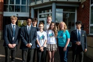 A group of seven secondary school students and an adult stand smiling outside a school building. Most students wear uniforms; two wear white T-shirts and a skirt, and the adult wears a blue “Buddy Leader” T-shirt.