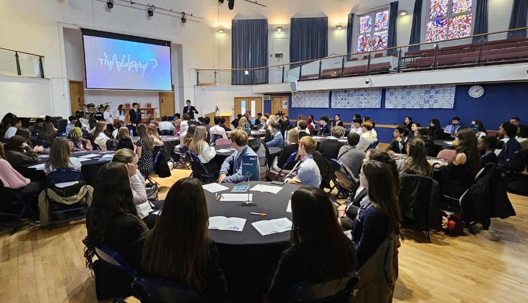 A large group of people sit around round tables in a spacious hall, listening to a presentation. A screen at the front displays “The purpose?” and stained glass windows are visible to the right.