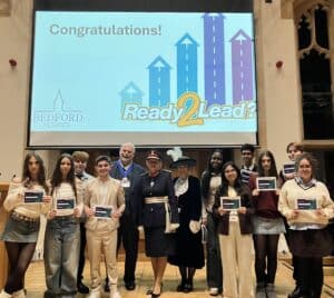 A group of students and adults stand on stage, proudly holding certificates. Behind them, a screen displays Congratulations!, the Bedford School logo, and the Ready 2 Lead? graphic—highlighting their impact as recent grant recipients.