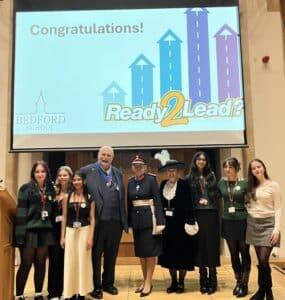 A group of nine people stand smiling on a stage in front of a screen that reads “Congratulations! Ready 2 Lead?” at Bedford School, celebrating the impact of a recent grant. The group includes adults in formal attire and young women.