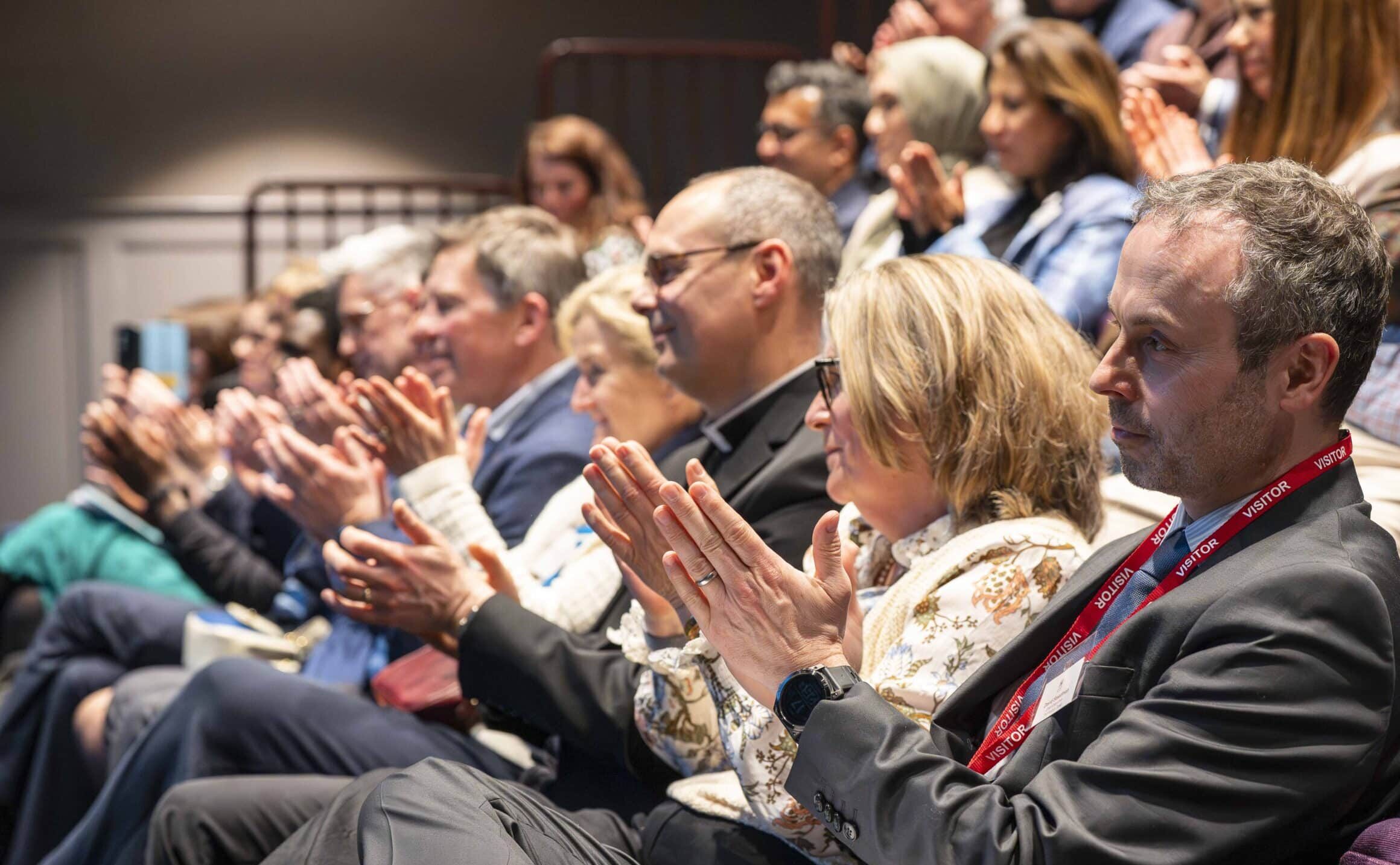 A group of people in theatre-style seats attentively watch an impactful event, all applauding. The audience members, both men and women in business attire, appear engaged with the presentation, appreciating its positive impact.