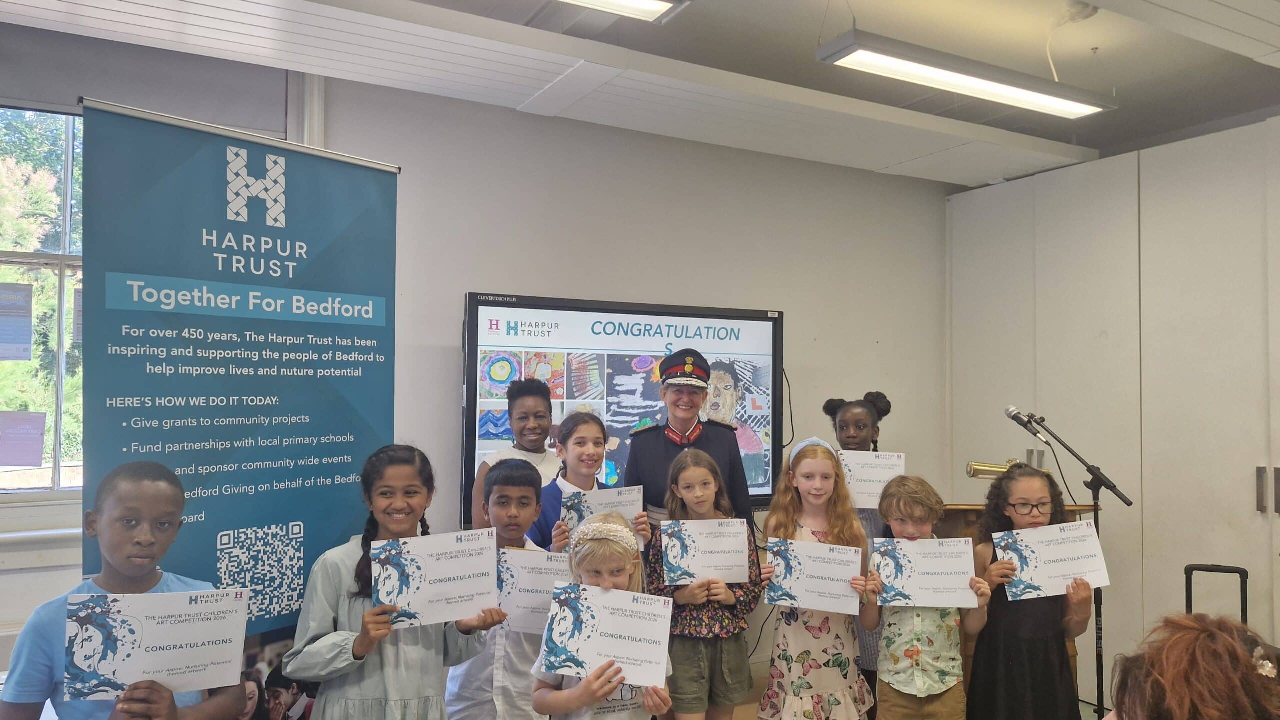A group of children stand in a classroom holding certificates, smiling at the camera. Behind them are two adults, a Harpur Trust banner, and a screen displaying CONGRATULATIONS, celebrating the impact of the grant on their achievements.