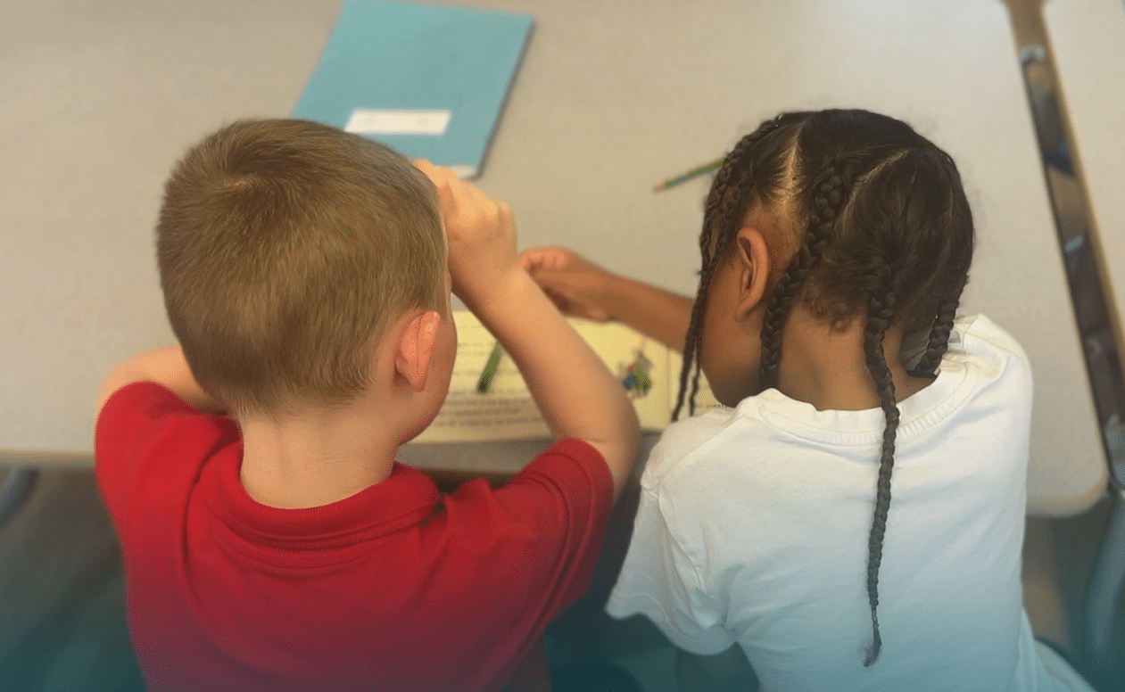 Two young children sit side by side at a table, working together on a worksheet or colouring activity. One wears a red shirt, the other a white shirt with plaited hair. Crayons and a folder are on the table.
