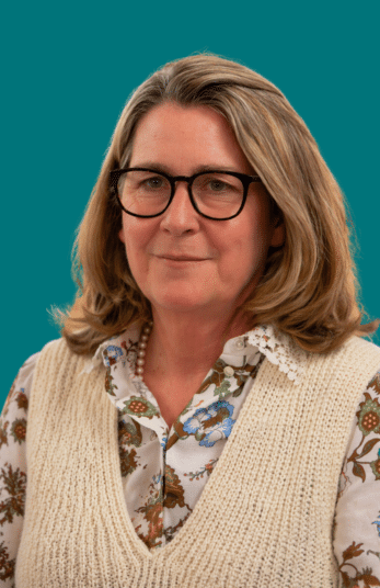 A Harpur Trust middle-aged woman with shoulder-length blonde hair and black-rimmed glasses, wearing a floral blouse under a cream knitted waistcoat.