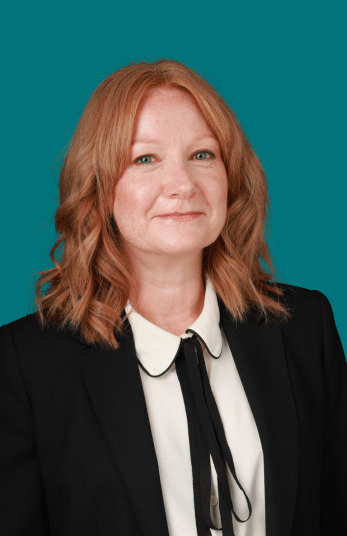 A Harpur Trust staff member with shoulder-length auburn hair, wearing a black blazer over a white collared shirt with a black tie.